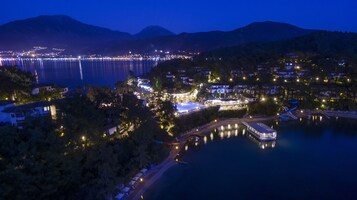 A coastal town at night with a marina, illuminated buildings, and a mountain backdrop.