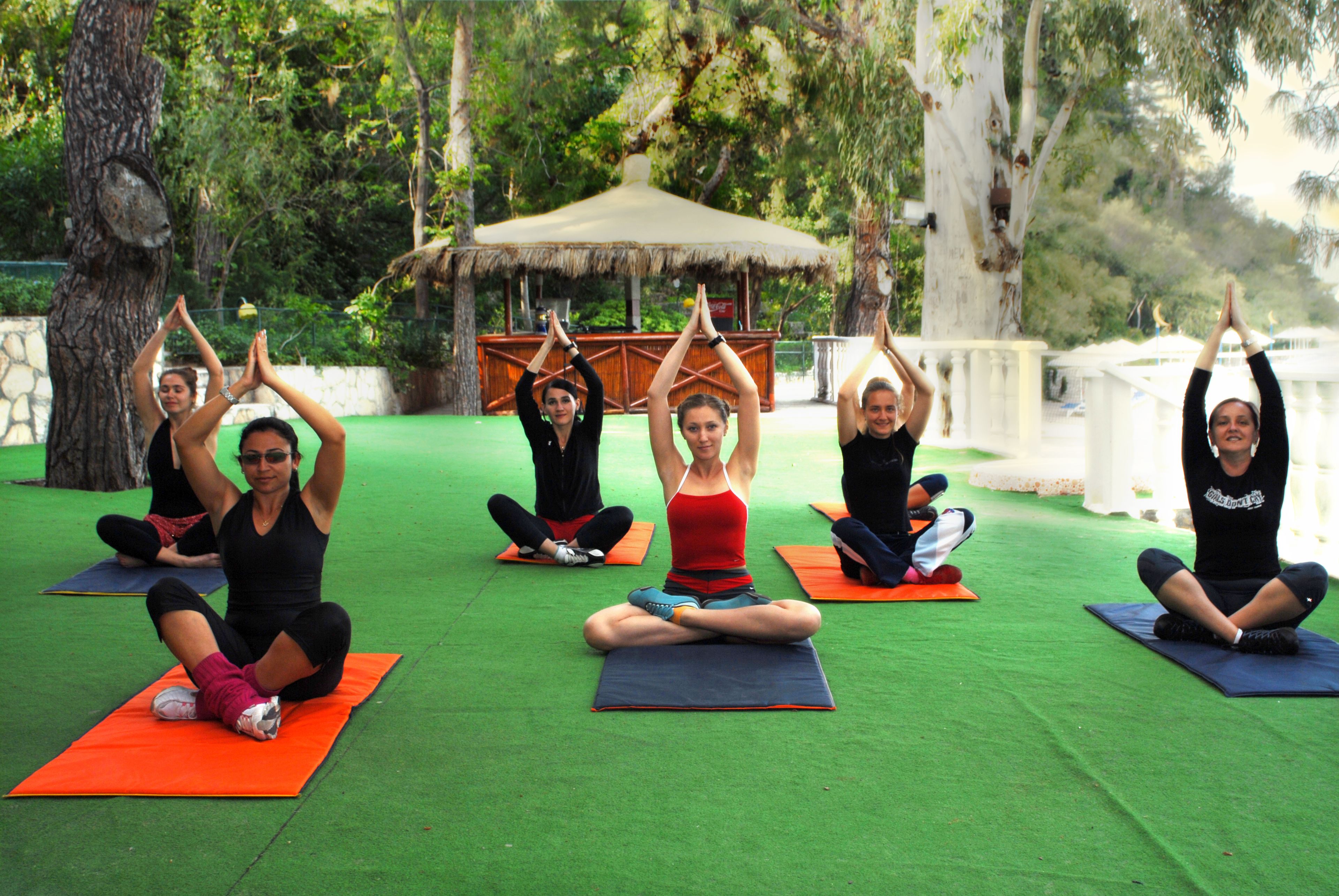 A group of people performing yoga on mats outdoors with trees and a thatched-roof structure in the background.