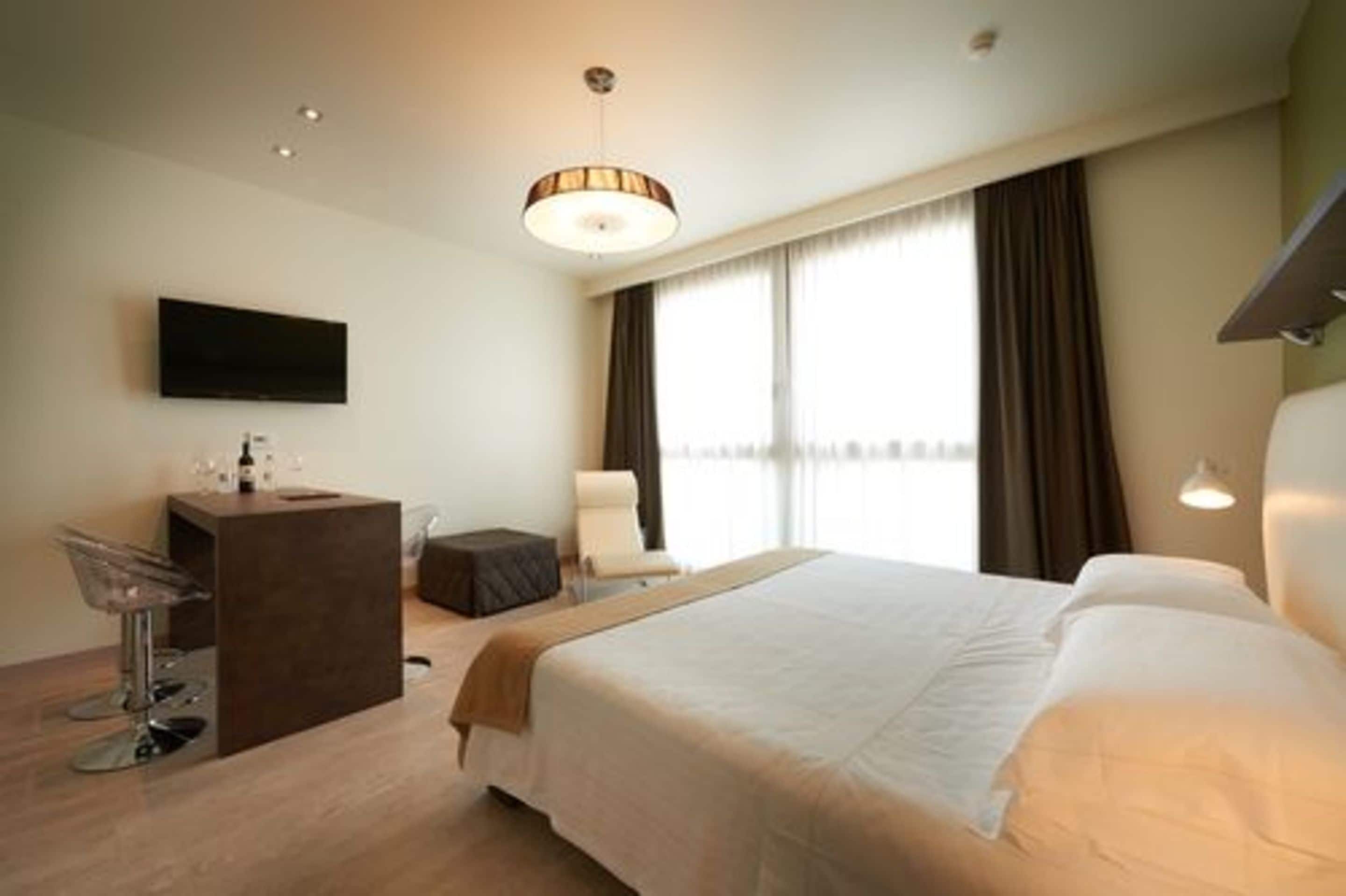 A hotel room with a bed, a desk with a chair, a TV, and a window with curtains.