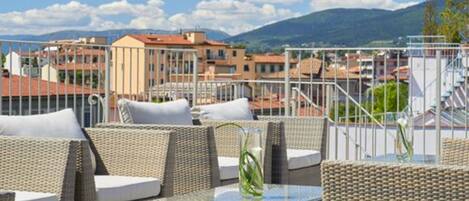 A terrace with wicker furniture and a glass table, overlooking a cityscape with buildings and mountains.