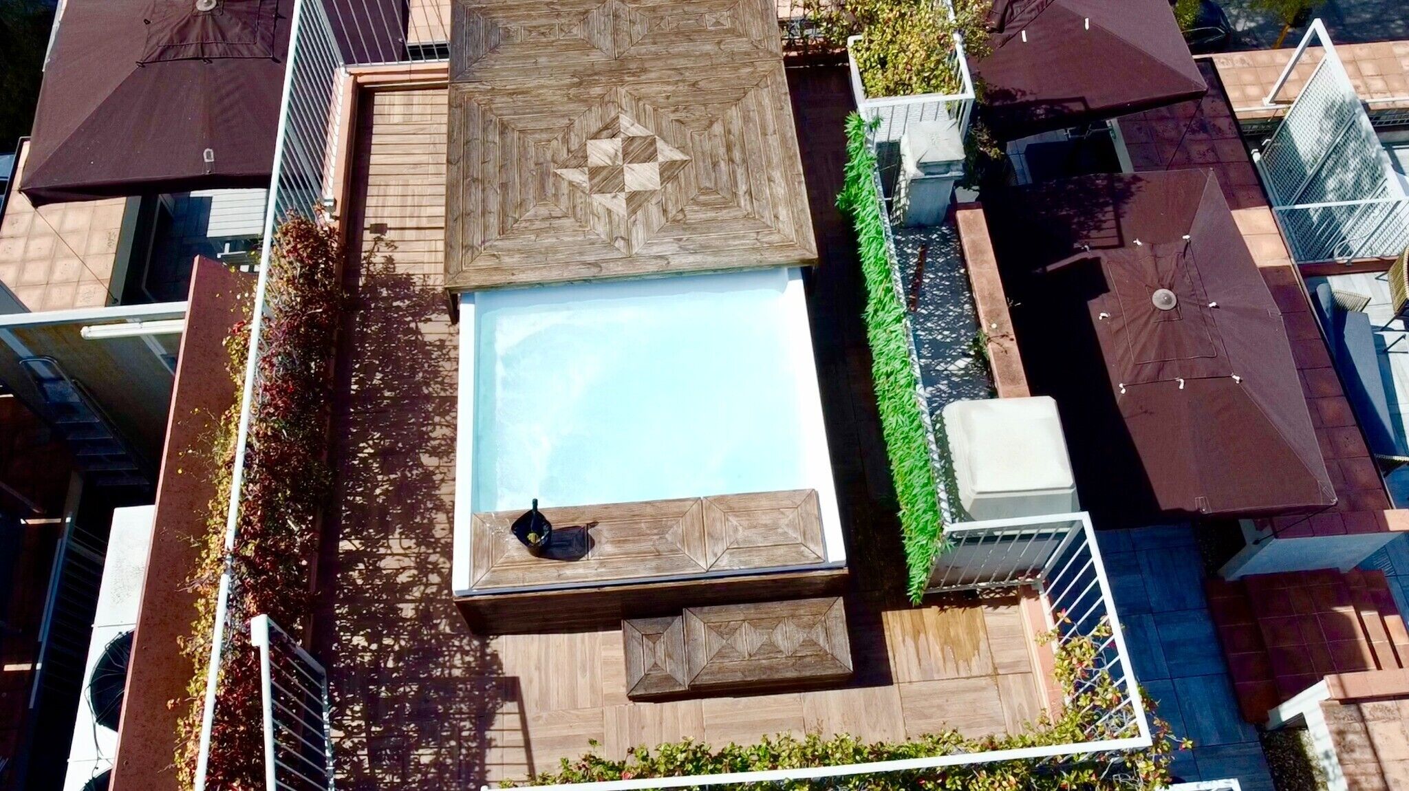 A rooftop pool area with a geometric wooden deck, surrounded by greenery and shaded by umbrellas.