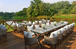An outdoor dining area with tables set for a meal, surrounded by chairs with white cushions, overlooking a body of water and greenery. - Selectum Family Resort Belek (Serik)
