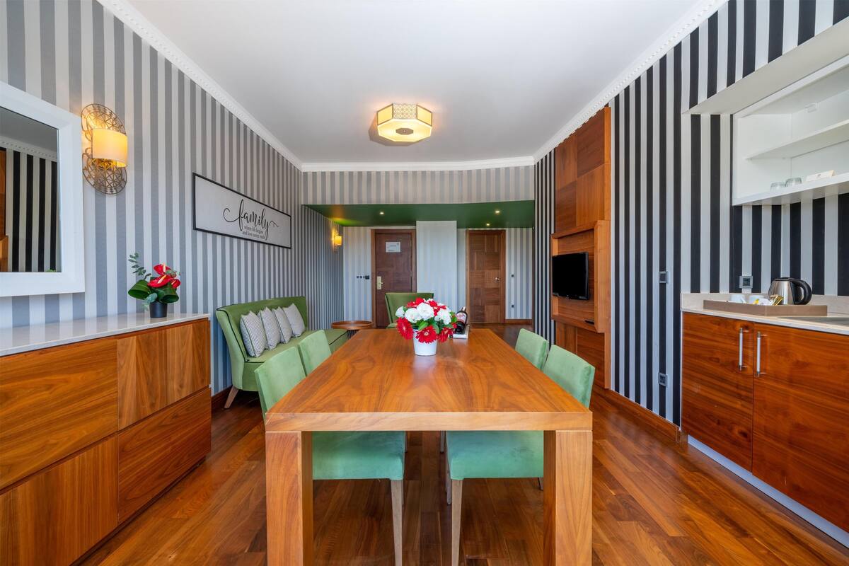 Un espace repas moderne avec une table en bois, des chaises recouvertes d’un tissu vert et un vase rempli de fleurs.