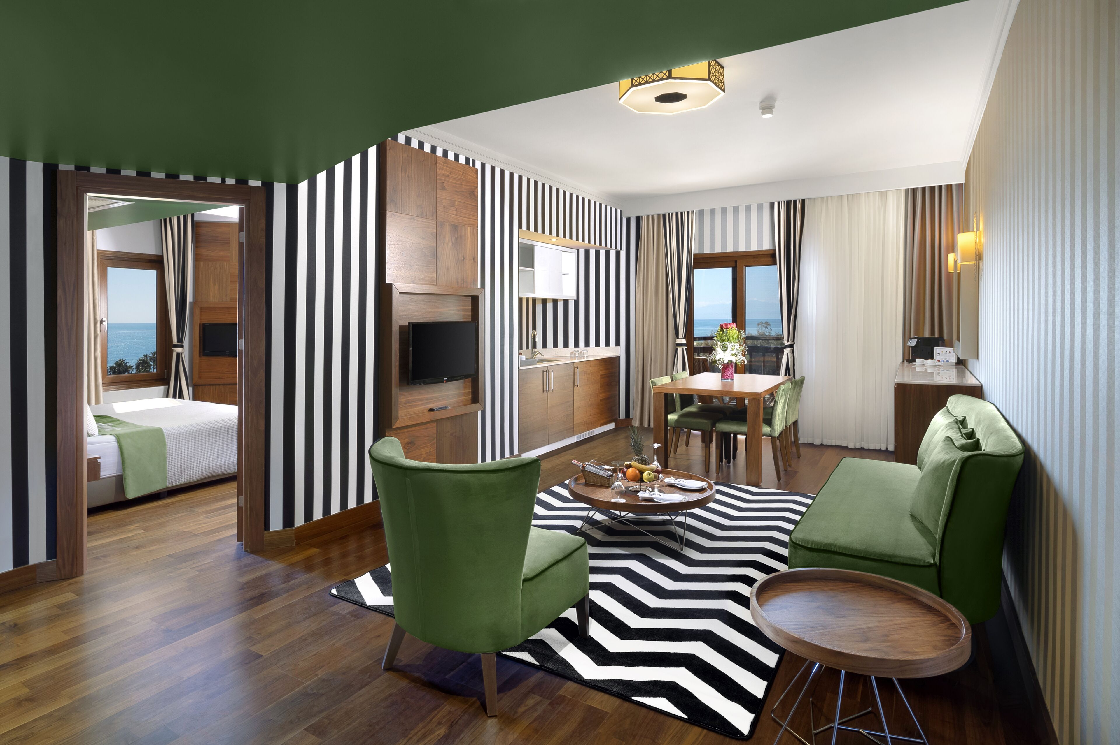 A modern hotel room with a bed, green armchairs, a dining area, and a view of the sea.