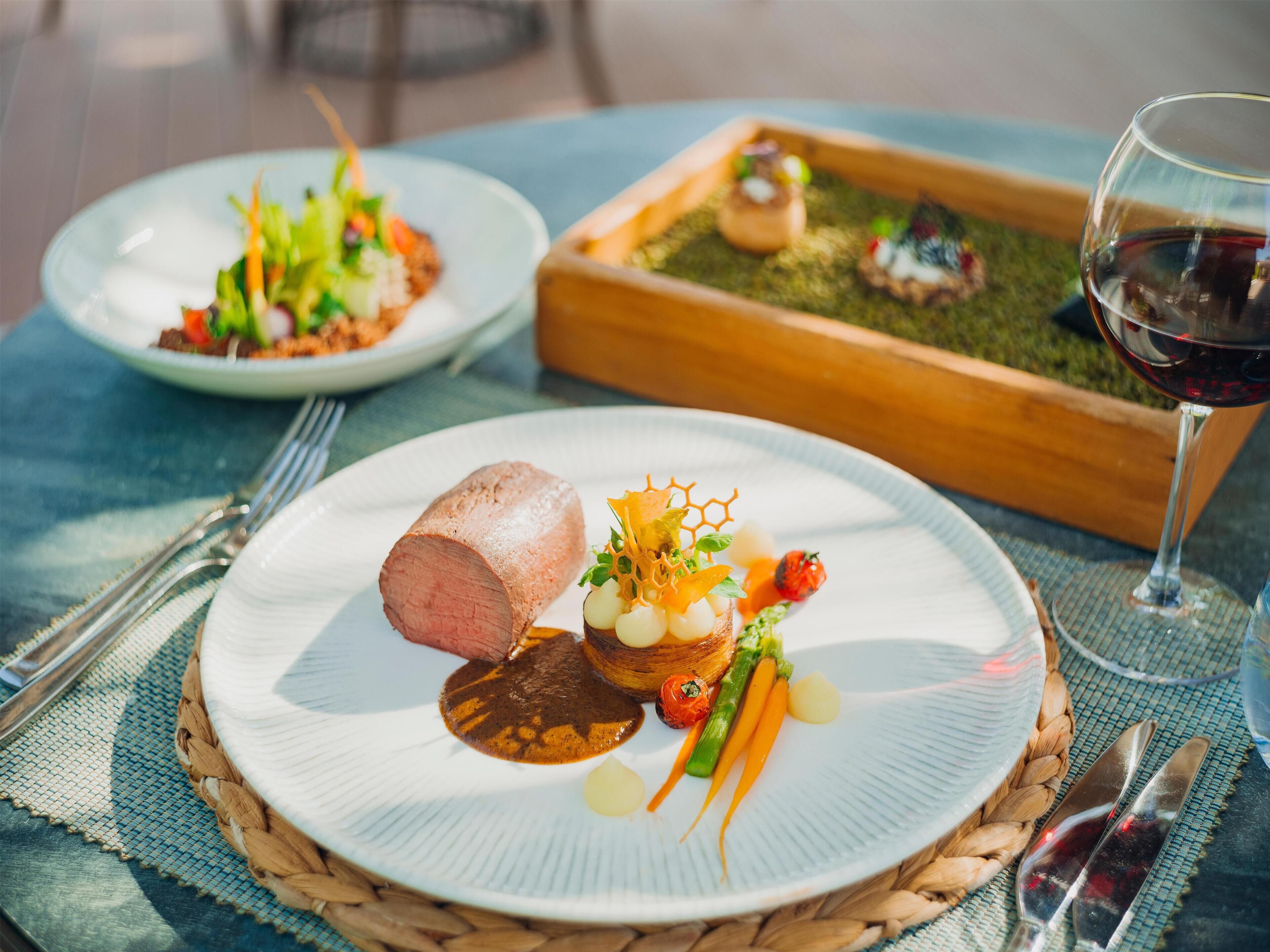 A dining table with plates of food, a glass of red wine, and a woven placemat.