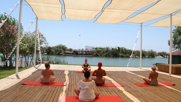 Un grupo de personas sentadas sobre esterillas de yoga en una terraza de madera, con vista a un río y un barco al fondo.