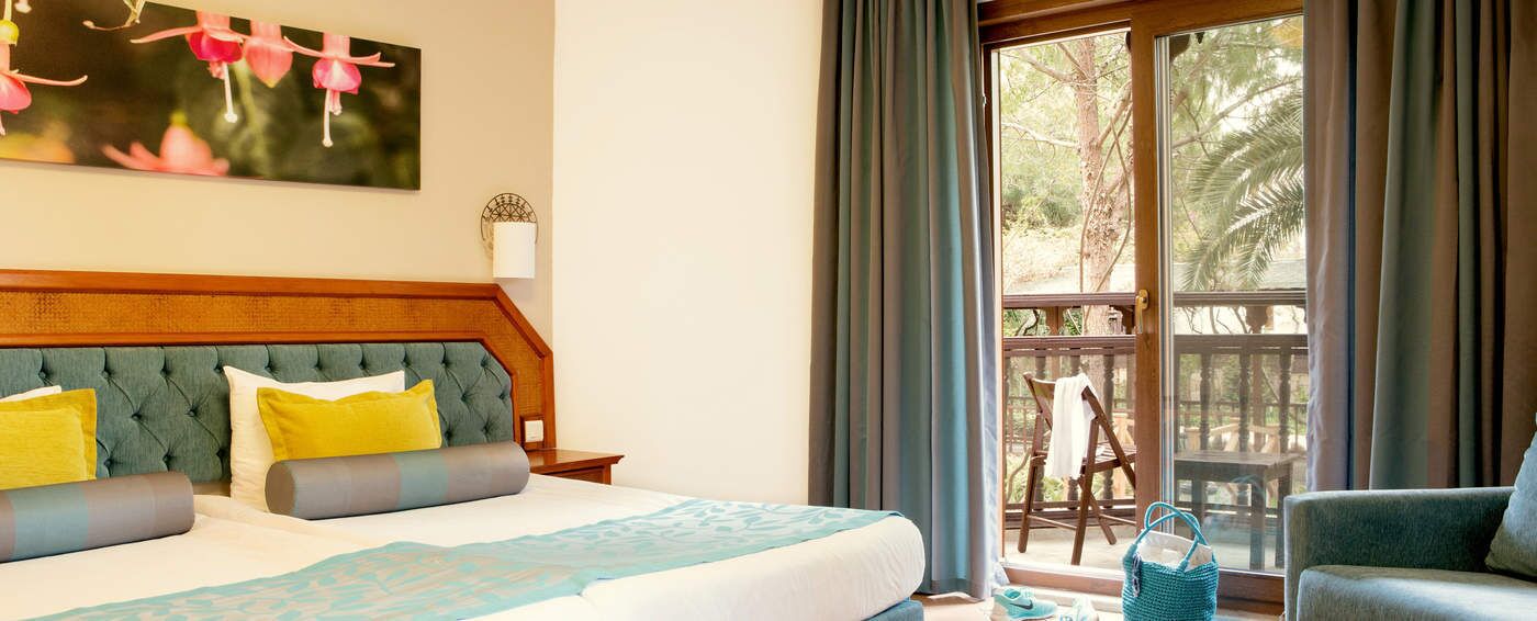 A hotel room with a large bed, a wooden headboard, a balcony with a view of trees, and a chair.