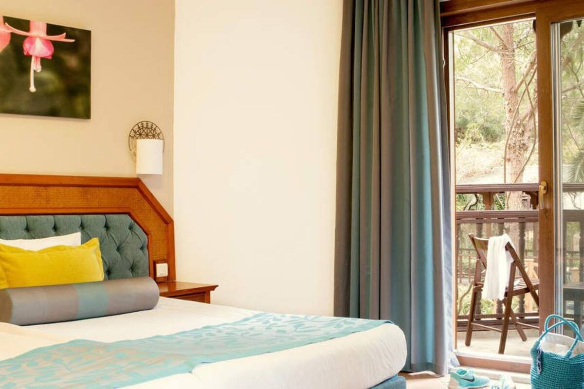 Une chambre d’hôtel avec un grand lit, une tête de lit en bois, un balcon donnant sur la verdure et un fauteuil.