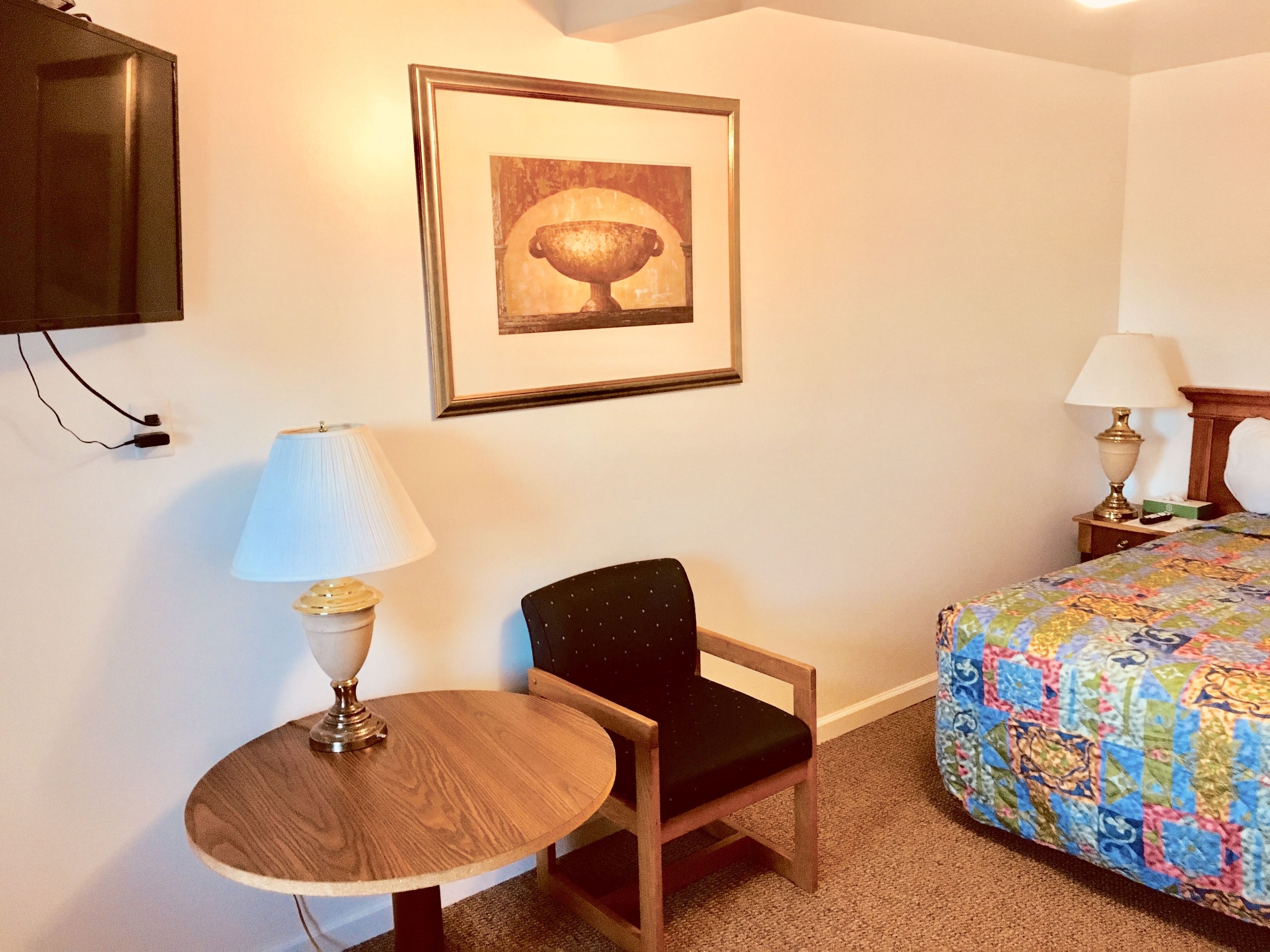Ein Hotelzimmer mit einem Bett, einem runden Holztisch mit Lampe, einem Sessel, einem gerahmten Bild an der Wand und einem an der Wand befestigten Fernseher.