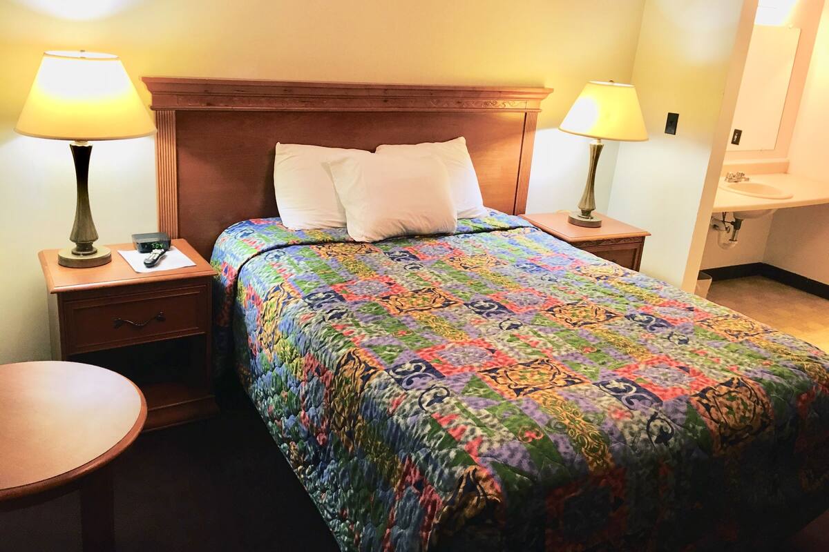 A hotel room with a wooden headboard, a colorful quilt, two bedside lamps, a bedside table, and a round table.