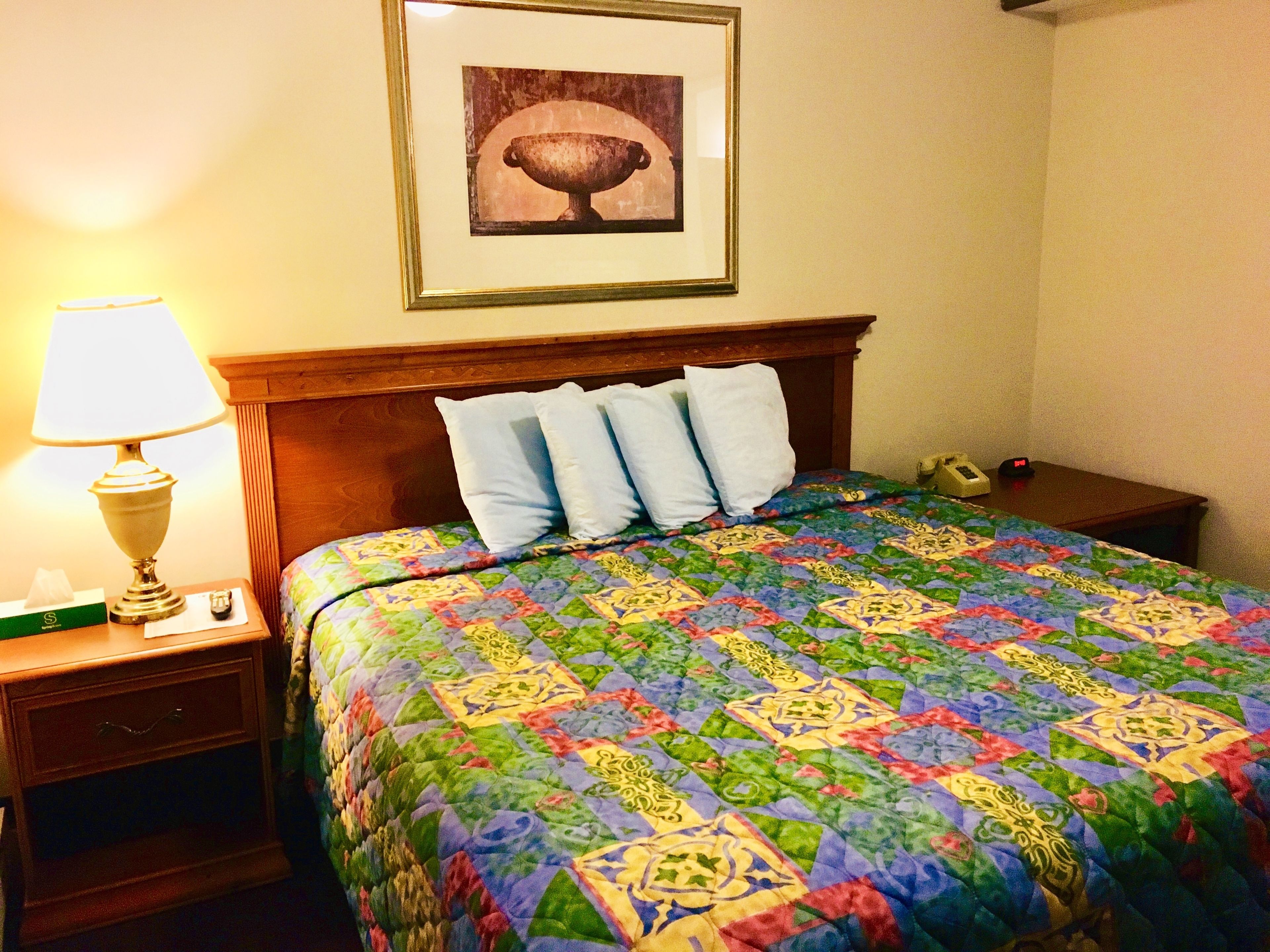 A hotel room with a wooden headboard, a colorful quilted bedspread, a bedside lamp, a framed picture on the wall, and a nightstand with a telephone and a tissue box.