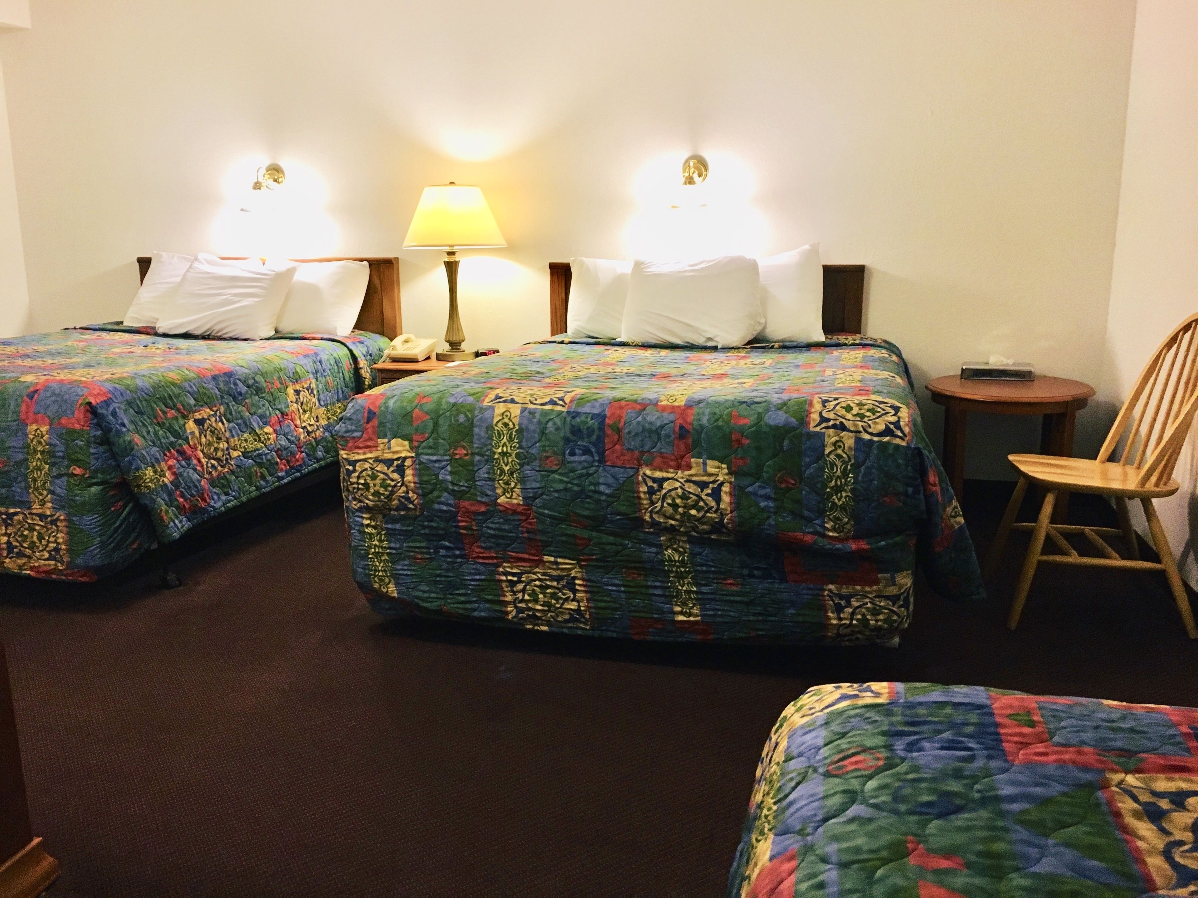 Hotellihuone, jossa on kaksi sänkyä. Jokaisessa sängyssä on värikäs peitto, yöpöydällä on lamppu ja huoneessa on myös tuoli.
