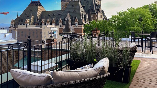 Una terraza en la azotea con mobiliario de mimbre, plantas en macetas y vistas a un edificio histórico con numerosas torres.