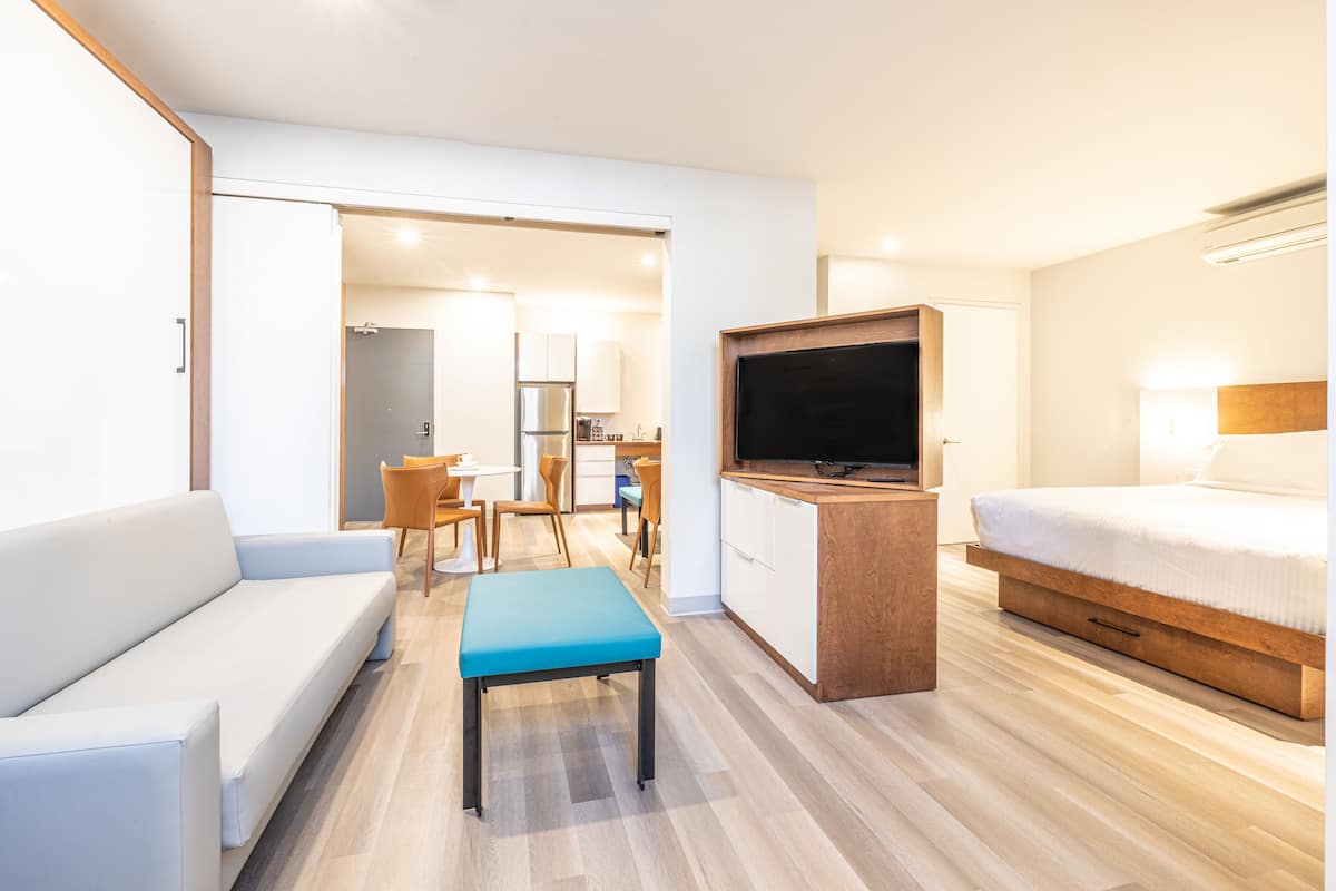 Ein modernes Hotelzimmer mit einer Couch, einem kleinen Tisch, einem Fernseher, einem Bett und einer Küchenzeile im Hintergrund.