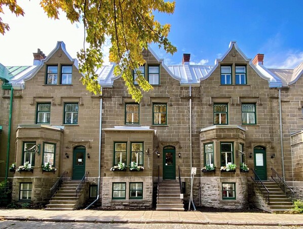 Una hilera de casas urbanas de piedra de tres pisos, con puertas y ventanas verdes, una entrada central con escalones, y un cielo azul claro arriba.