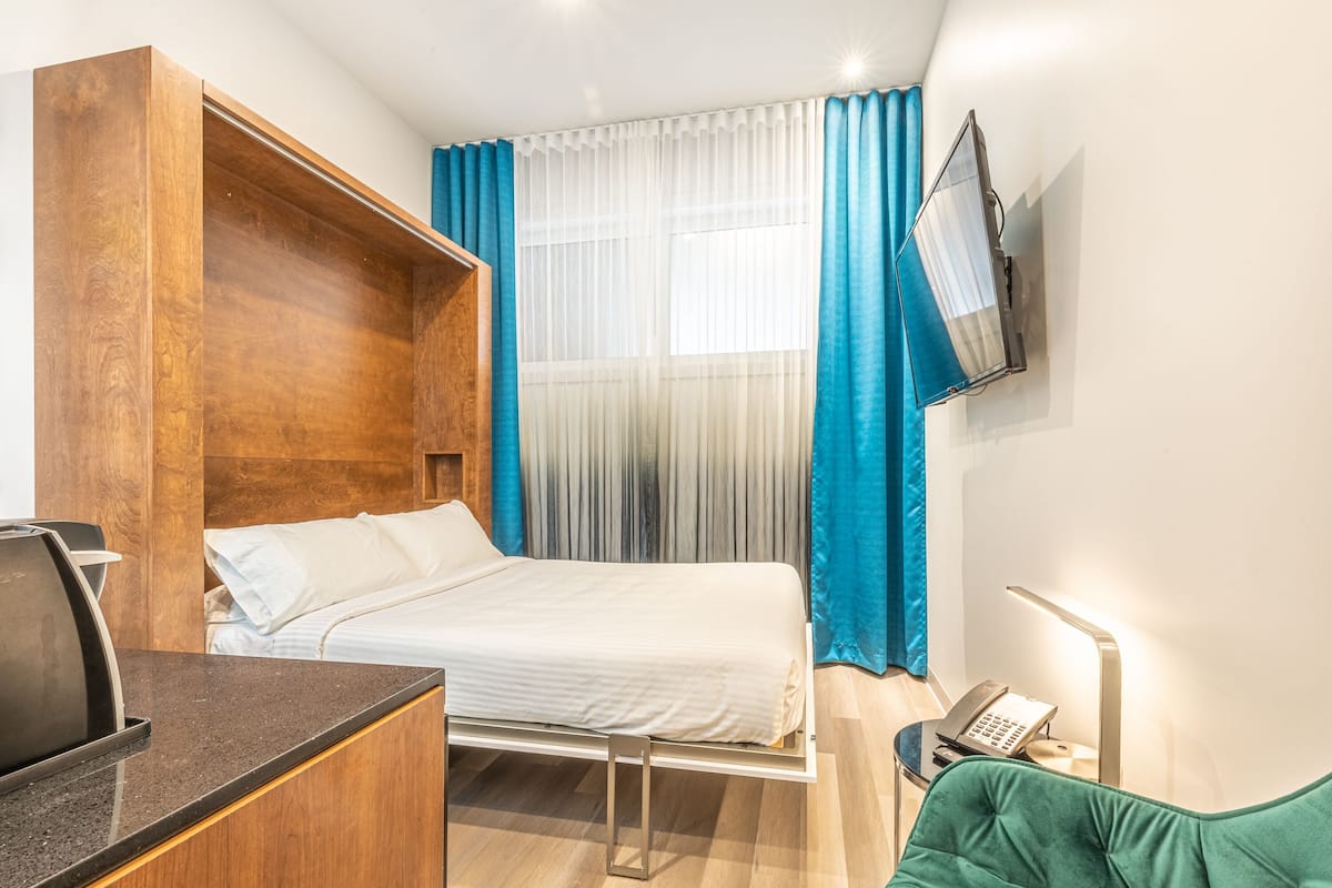 Ein Hotelzimmer mit Bett, Schreibtisch, Stuhl, an der Wand montiertem Fernseher und Nachttisch mit Lampe.