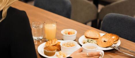 Una persona sentada a una mesa de madera con un plato que incluye un muffin, un croissant y una bebida, con sillas y una mesa al fondo.