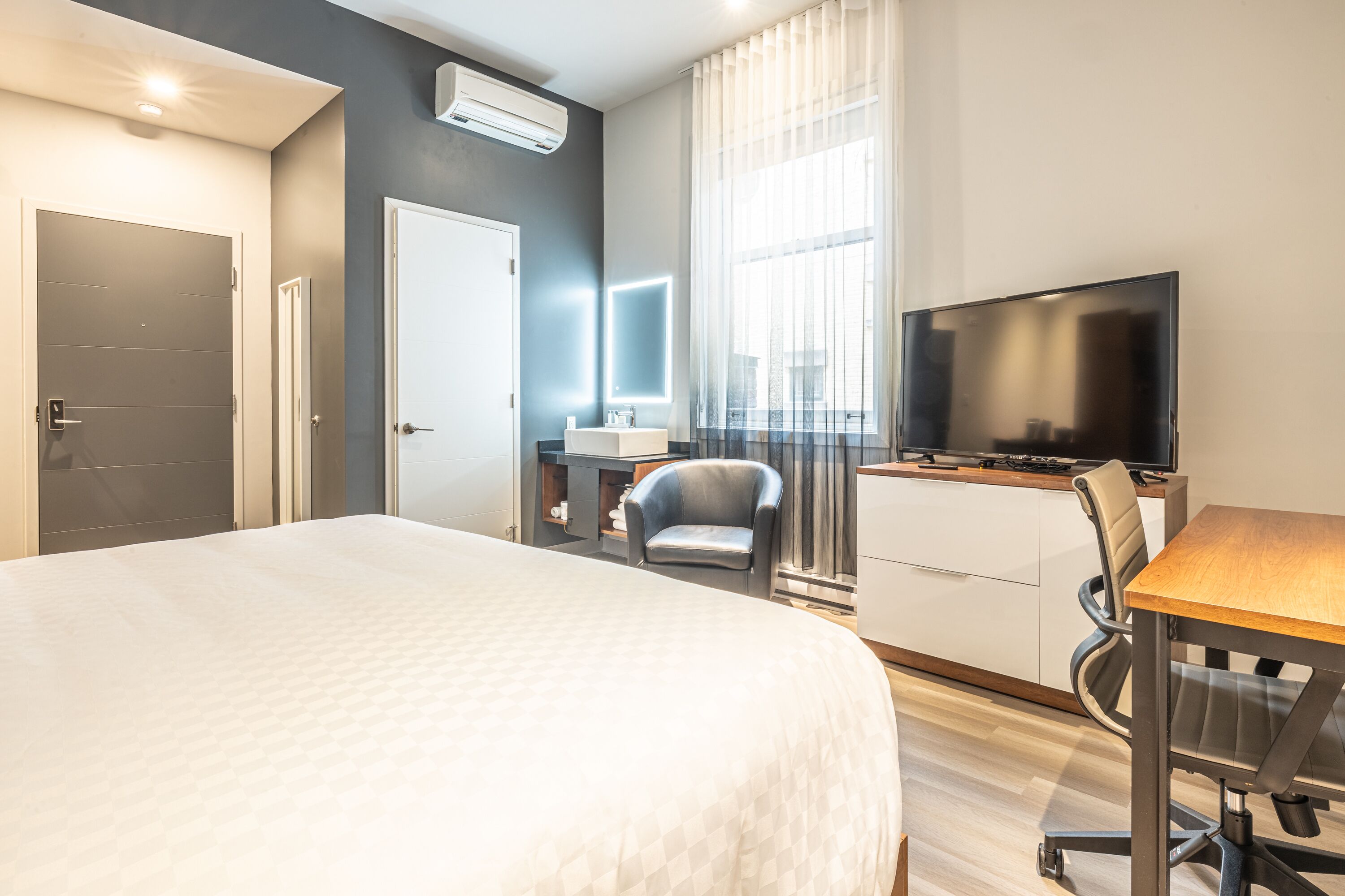 A hotel room with a large bed, a desk with a chair, a TV, a small table, and a window with curtains.