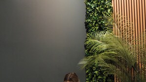 Una persona sentada junto a una piscina, con una toalla, rodeada de plantas de interior y un panel de madera.