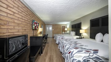 A hotel room with a brick wall, a television, a desk, a bed with patterned bedding, and two bedside lamps.