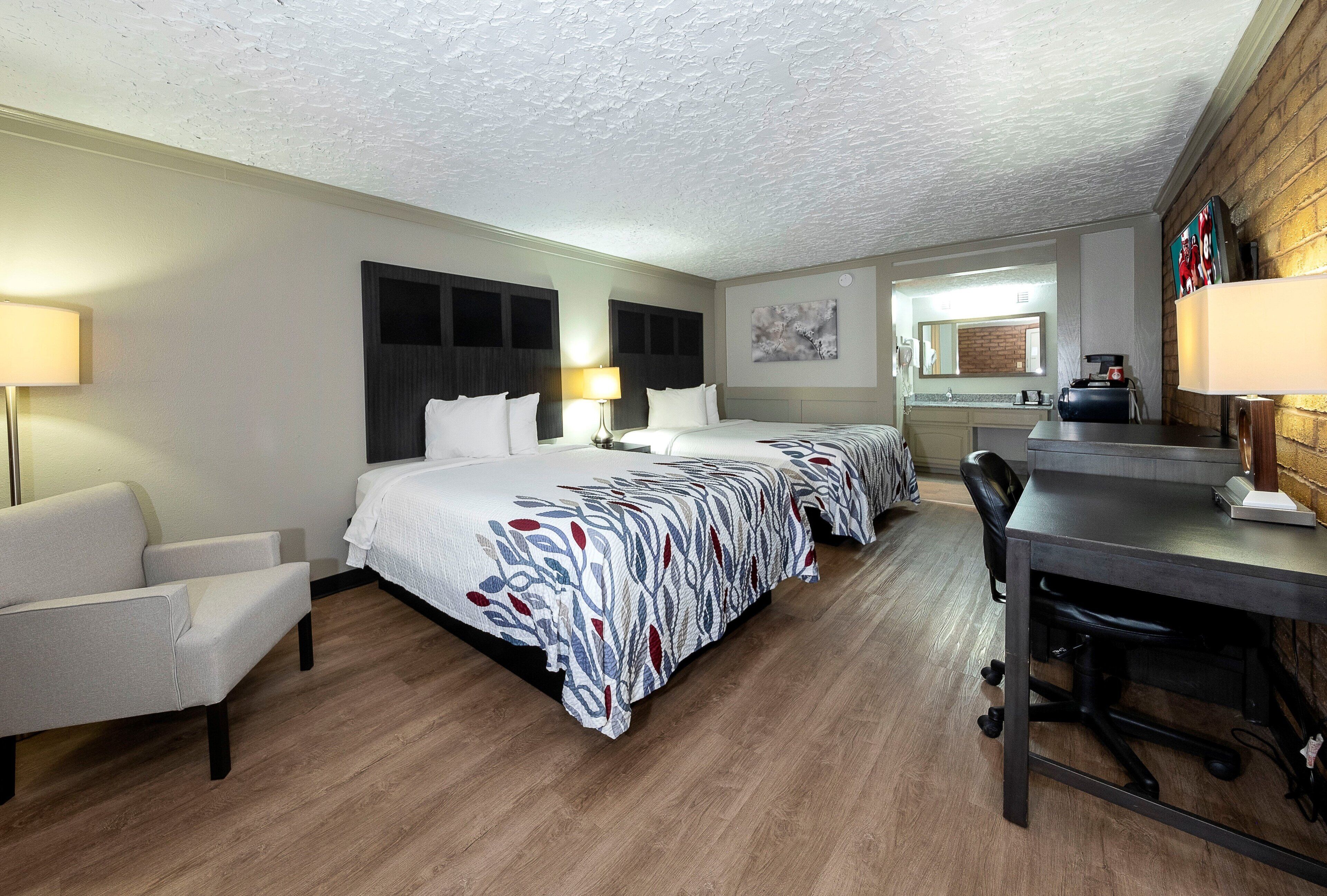 A hotel room with a large bed, two bedside tables, a desk with a chair, a lamp, a television, and a bathroom visible through an open door.