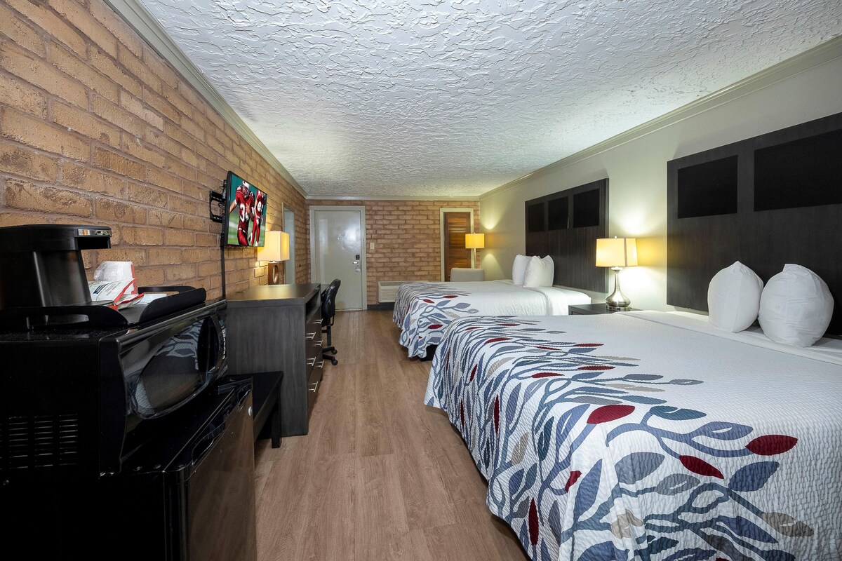 A hotel room with a brick wall, a flat-screen TV, a desk with a coffee maker, two beds with patterned bedspreads, and two bedside lamps.