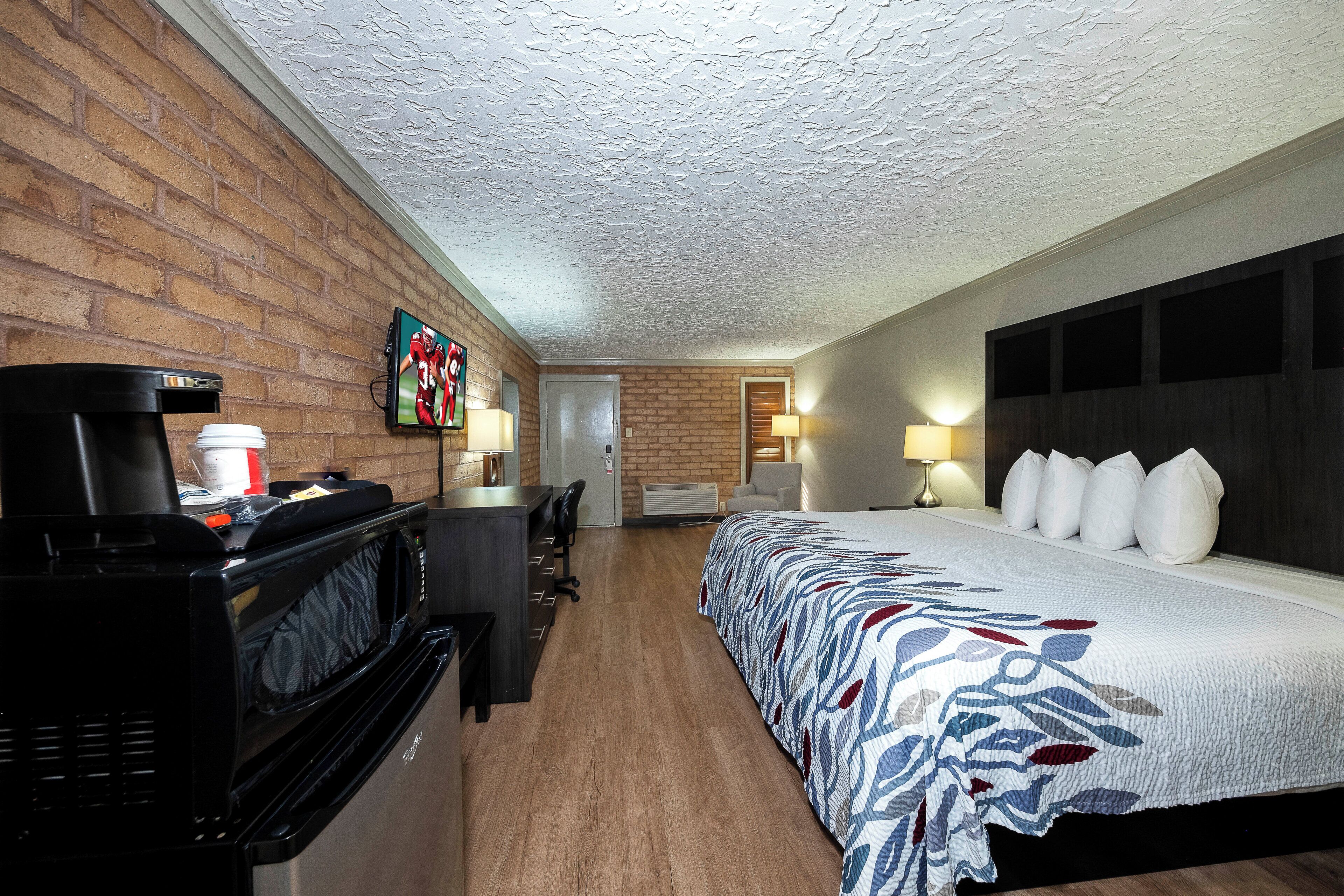 A hotel room with a brick wall, a bed with a patterned bedspread, a desk with a coffee maker, a television, and a door leading to another room.