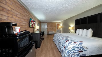 A hotel room with a brick wall, a bed with a patterned bedspread, a desk with a coffee maker, a television, and a door leading to another room.