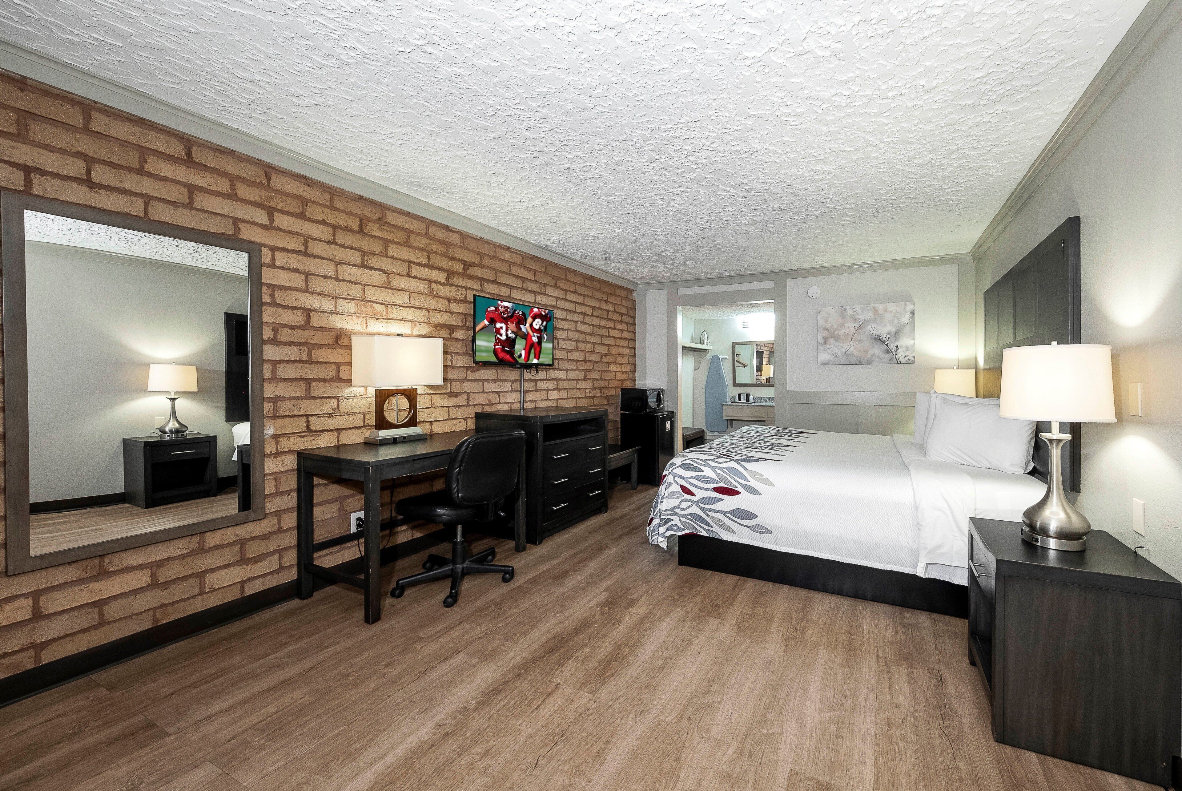 A hotel room with a brick accent wall, a bed with a patterned bedspread, a desk with a chair, a television, and a large mirror.