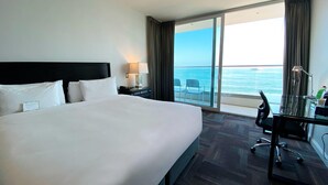 Habitación de hotel con una cama grande, un escritorio con silla y vista al océano a través de una puerta corrediza.