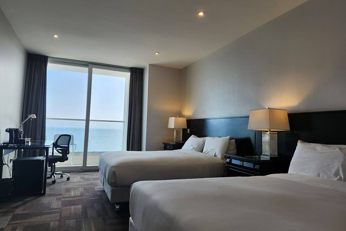 Une chambre d’hôtel avec deux lits, un bureau avec une chaise, une lampe et une vue sur la mer.