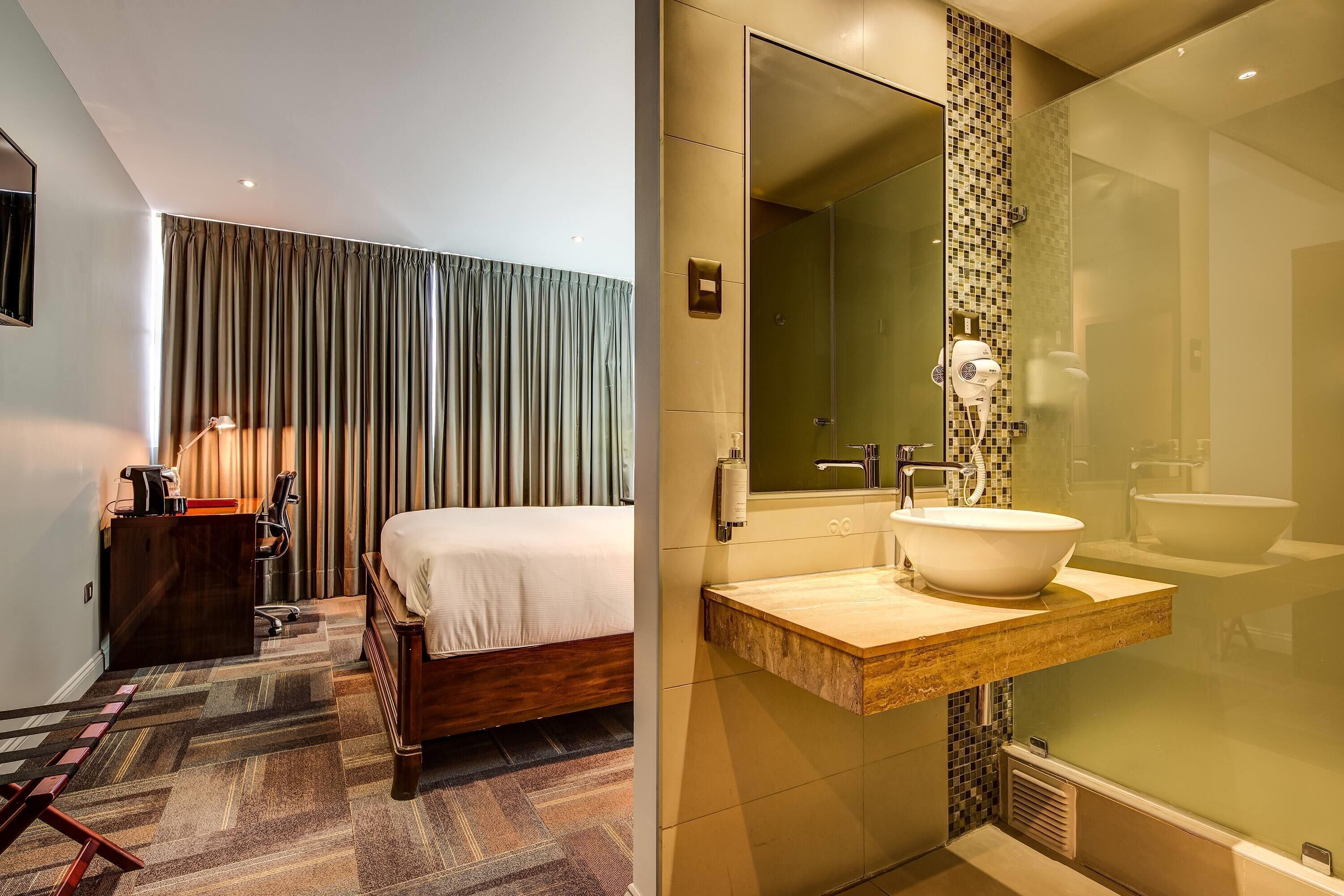 A hotel room with a bed, desk, and a bathroom with a large mirror, sink, and shower.