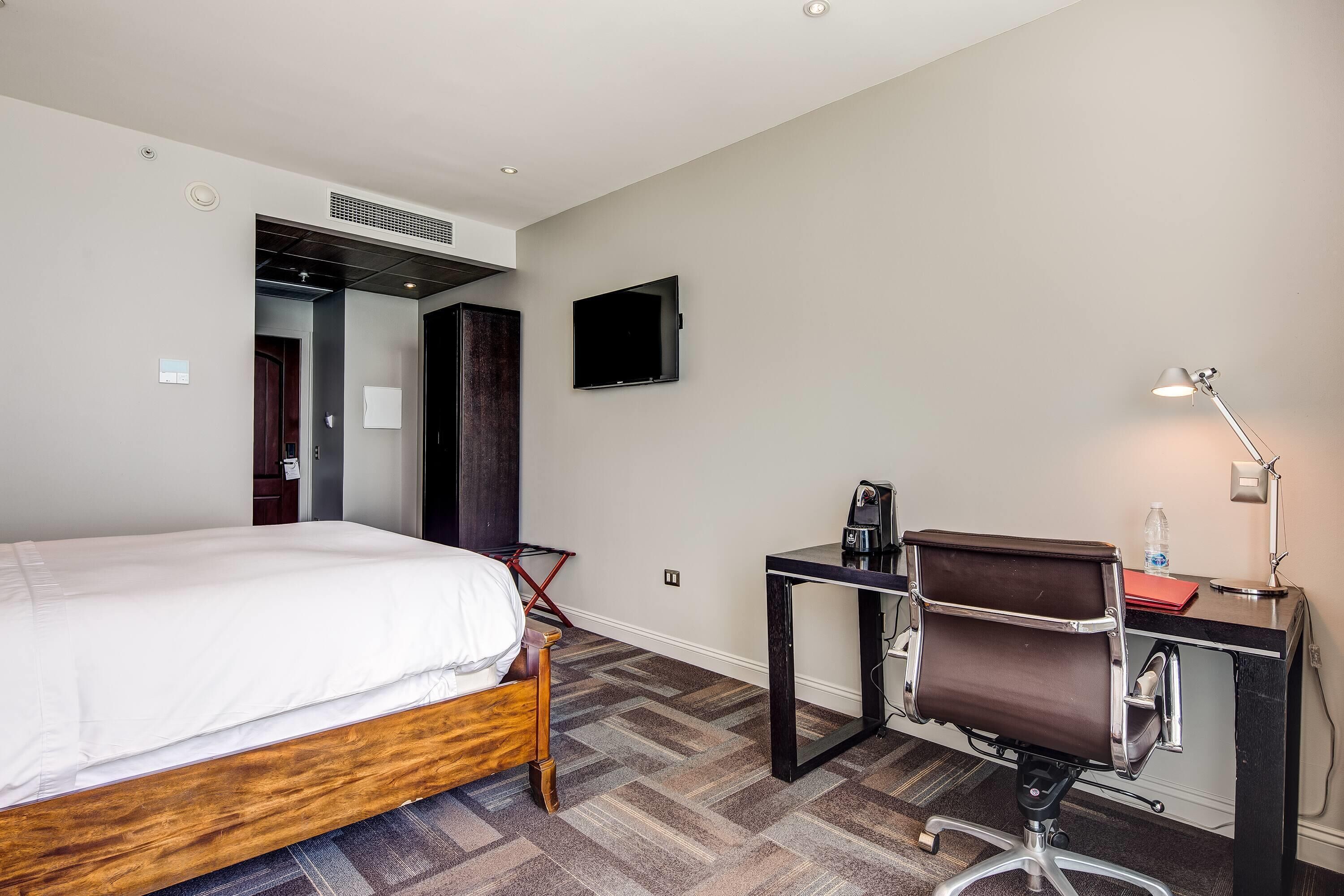 A hotel room with a bed, desk, chair, and a TV on the wall.