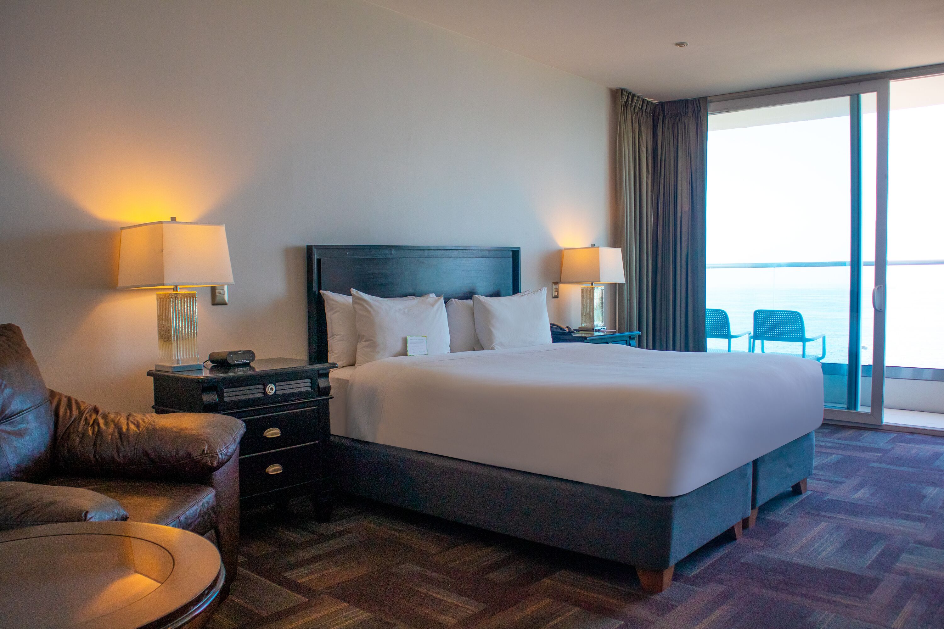A hotel room with a large bed, two bedside tables with lamps, a leather armchair, and a view of the sea through a sliding glass door.