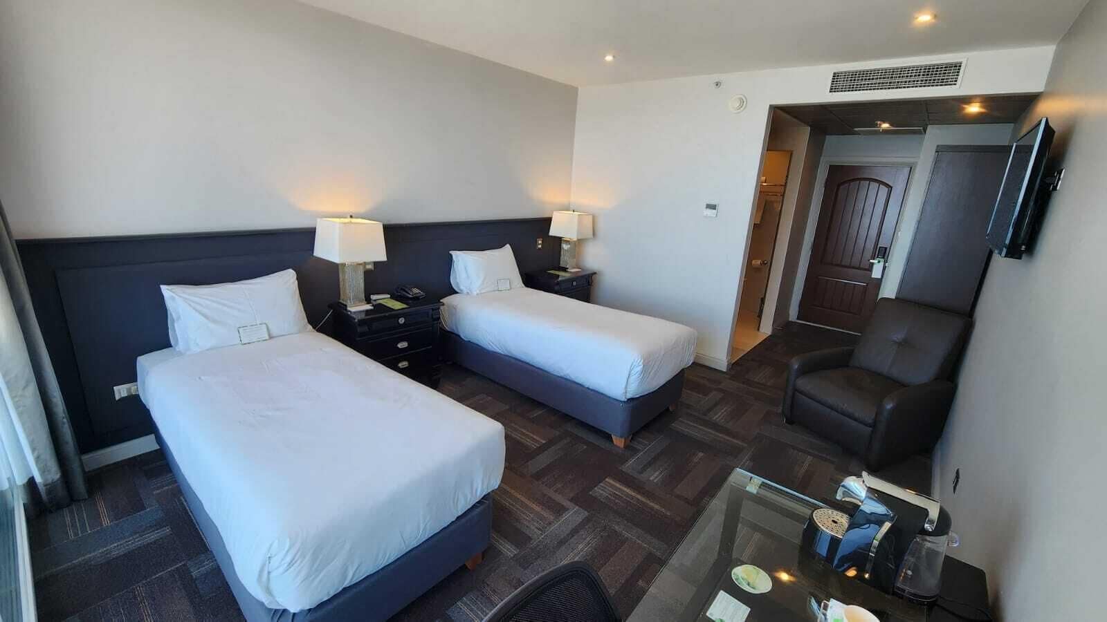 Habitación de hotel con dos camas, un escritorio con silla, televisión y puerta de acceso a un baño.