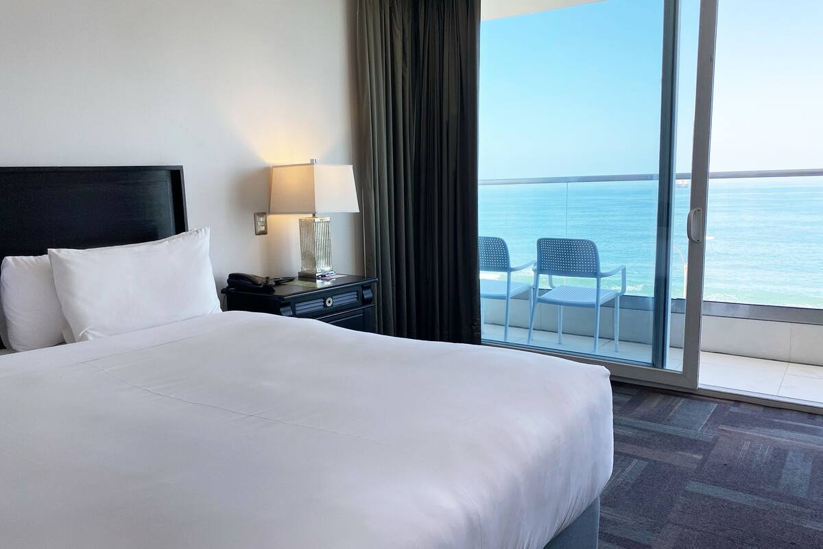Une chambre d’hôtel avec un grand lit, une table de chevet avec une lampe et une vue sur la mer, accessible par une porte-fenêtre coulissante.