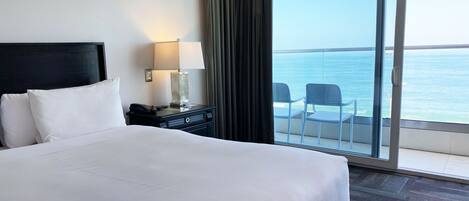 A hotel room with a large bed, a nightstand with a lamp, and a view of the sea through a sliding glass door.