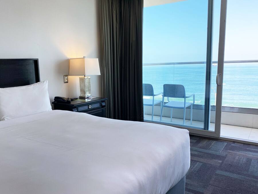 Habitación de hotel con una cama grande, una mesita de noche con lámpara y vista al mar a través de una puerta corrediza de vidrio.