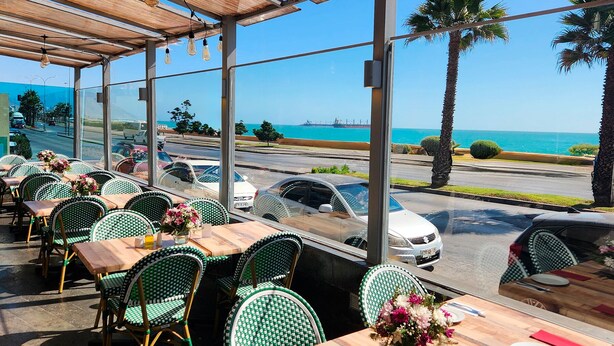 Un restaurante al aire libre con sillas de mimbre verde y mesas de madera, con vista a una carretera costera con coches y un cielo azul despejado.