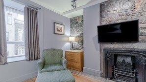 A living room with a green patterned armchair, a wooden dresser, a flat-screen TV, and a stone accent wall. - Hôtel Jardin Du Gouverneur (Québec City)