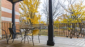 A wooden deck with two chairs and a table, a brick building in the background, and a metal railing. - Hôtel Jardin Du Gouverneur (Québec City)