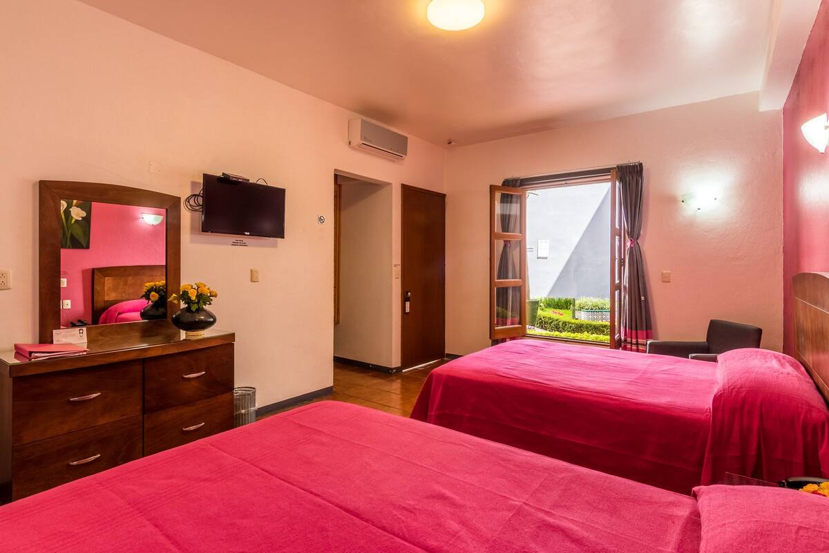 A hotel room with two beds, a TV, a desk with a mirror, and a window with a view of greenery.