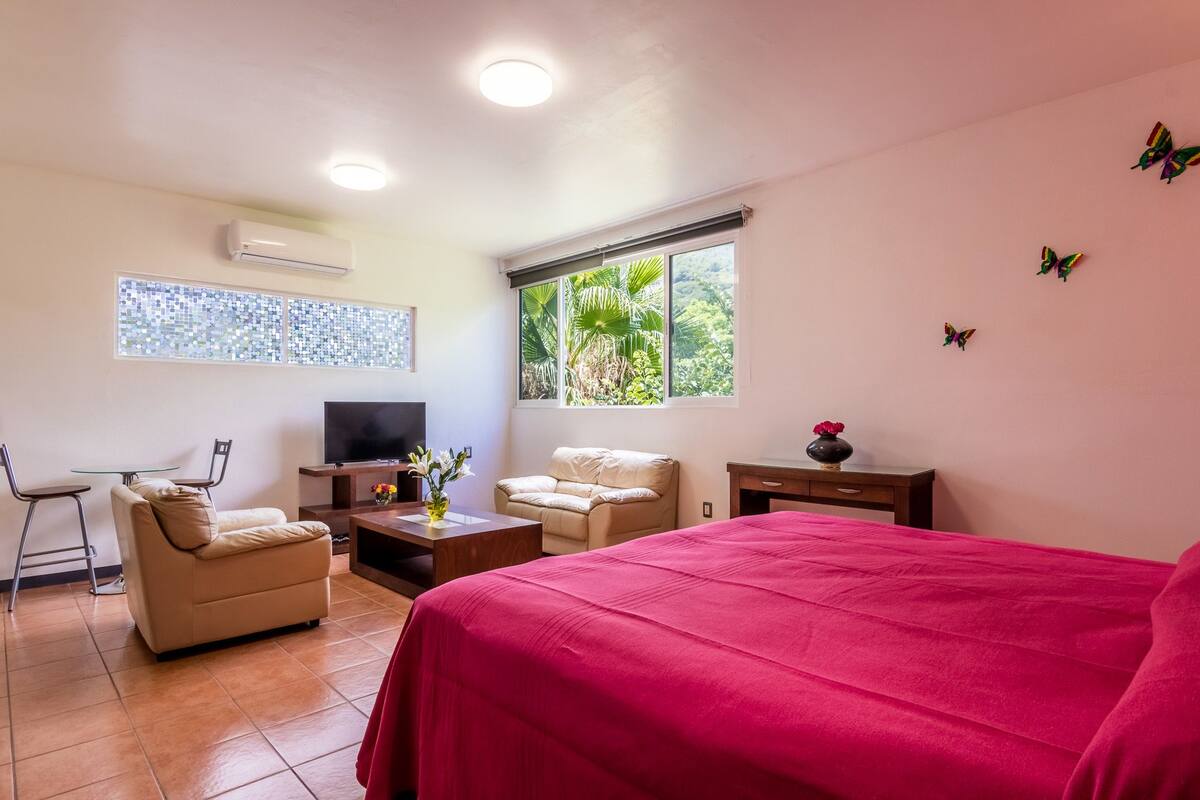 A hotel room with a bed, a TV, a sofa, a coffee table, and a window with a view of greenery.