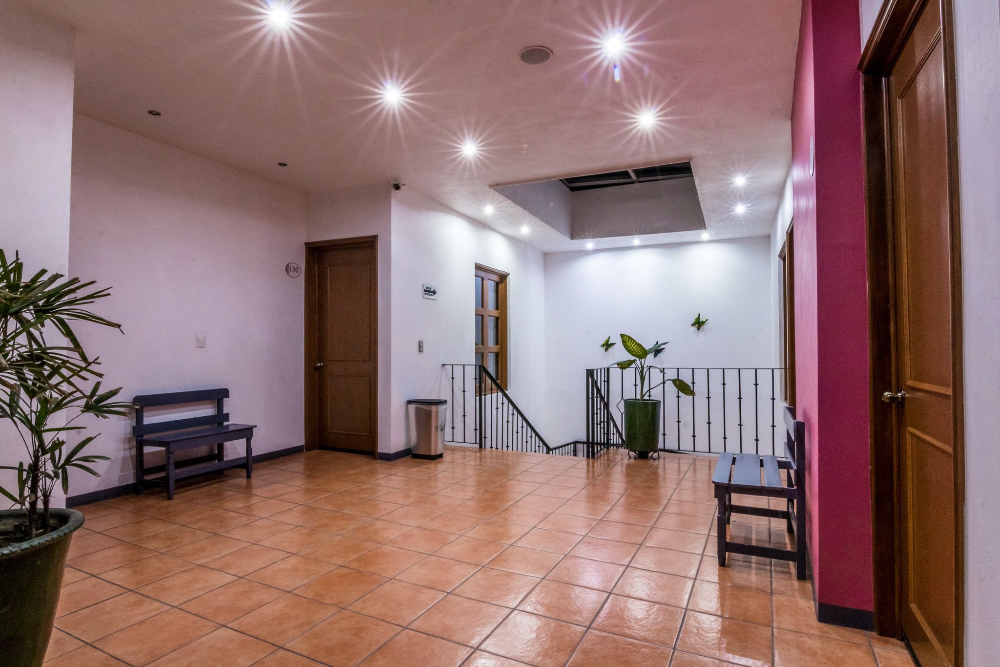 Um espaço amplo e coberto, com chão em piso cerâmico, um banco, um cesto de lixo e um vaso de planta.