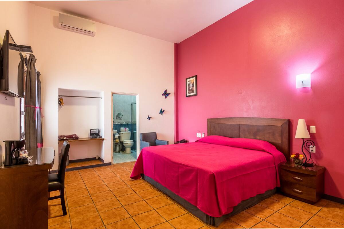 A hotel room with a bed, a desk, a chair, a nightstand, and a bathroom visible through an open door.