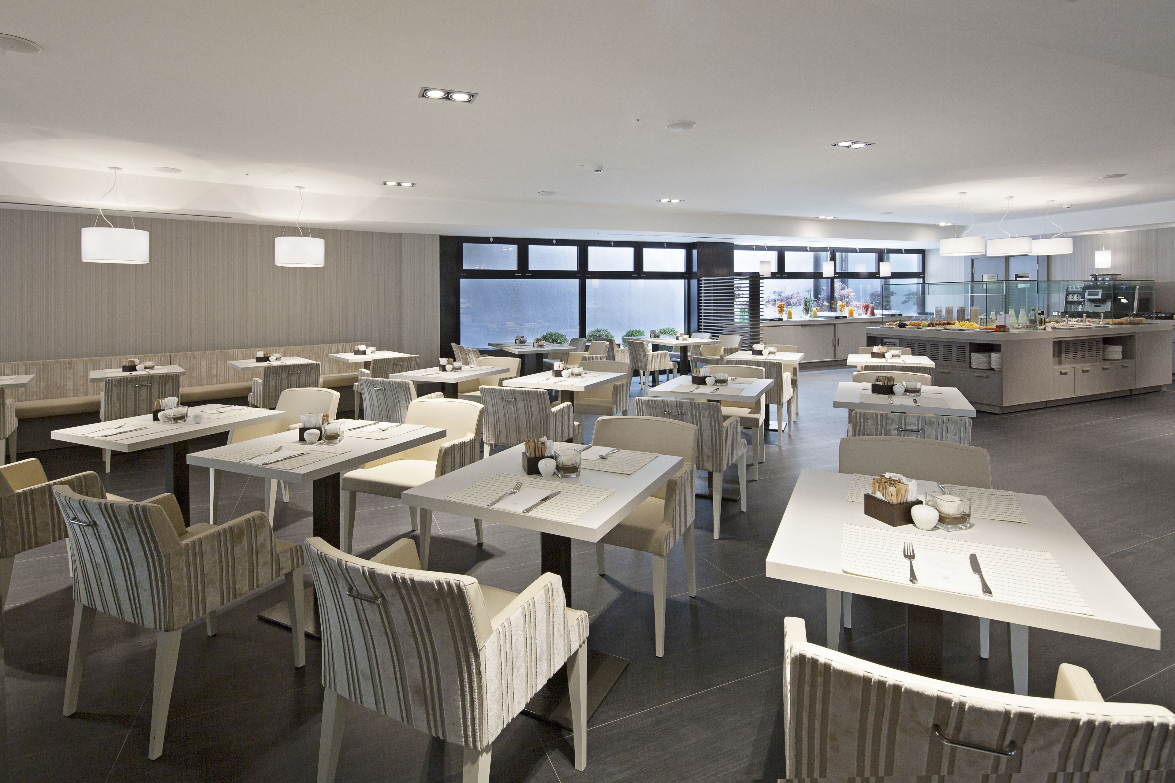 Un comedor moderno con varias mesas y sillas, ventanas amplias y una zona de cocina al fondo.