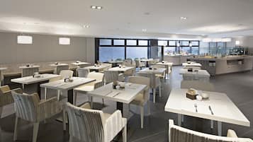Un comedor moderno con varias mesas y sillas, ventanas amplias y una zona de cocina al fondo.