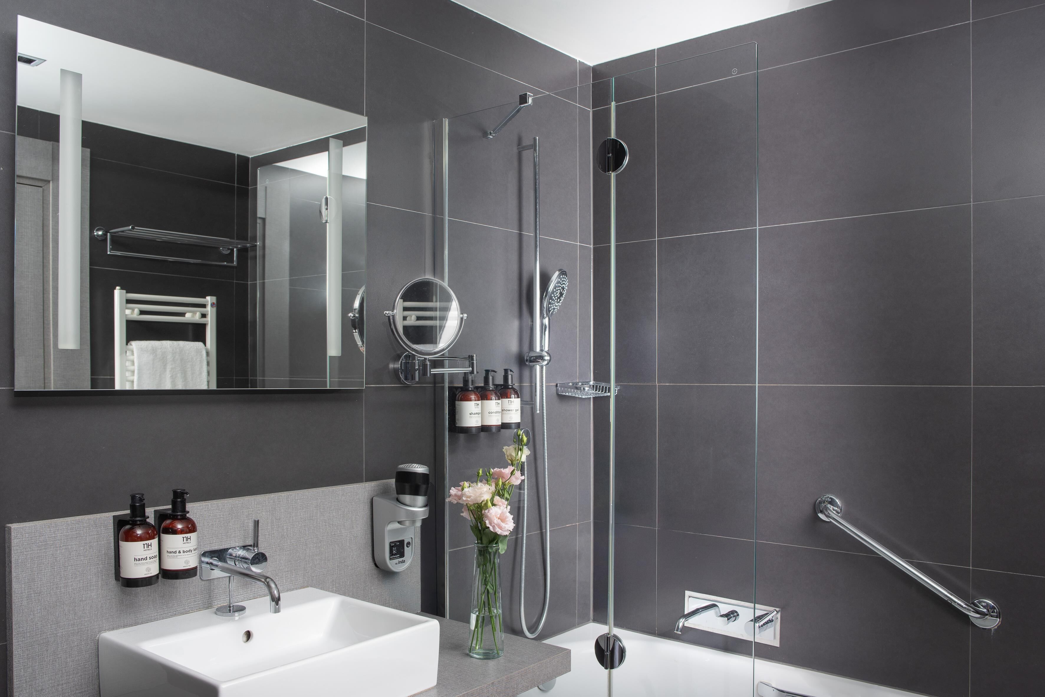 Un baño moderno con lavamanos blanco, espejo y ducha. Hay artículos de tocador en la pared y un florero con flores sobre el lavamanos.