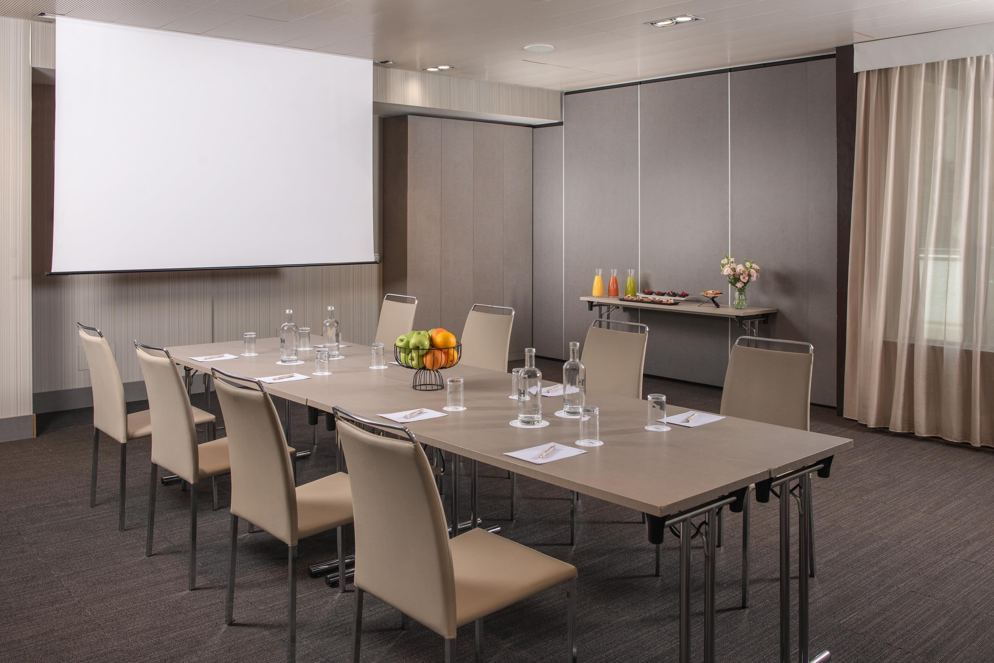 A conference room with a long table, chairs, a projector screen, and a small table with bottles and flowers.