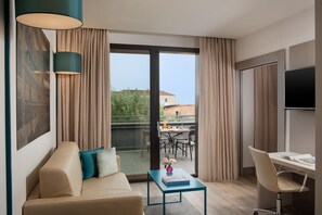 A modern hotel room with a sofa, a small table with a book, a desk with a chair, a TV, and a balcony with a table and chairs.
